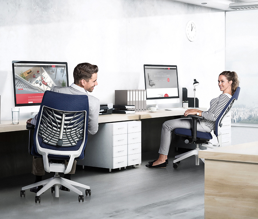 Sfeerbeeld op kantoor van een man en een vrouw op een donkerblauwe bureaustoel Joyce met FlexGrid en zwarte T-armleggers. Dankzij de in de bureaustoelen toegepaste Flextech-techniek zitten ze in een dynamische positie. De bureaustoel Joyce is van achteren én van opzij te zien.