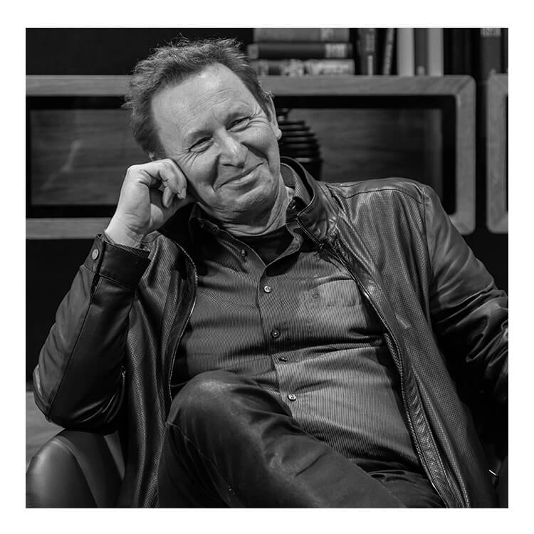 The designer of the SHUFFLEis1, Martin Ballendat, smiling and sitting on a chair, supporting his head with his hand. He is wearing jeans and a shirt with a leather jacket.