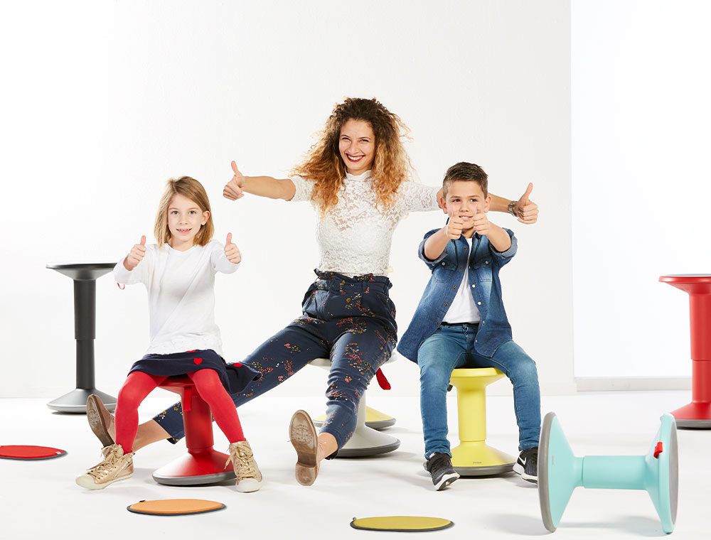 Two children and a women are sitting on colourful UP Junior stools. They are smiling and giving a thumbs up to the camera. On the ground, there are UP seat cushions in several colours (green, orange, red). In the background, there are two tall UP stools in black and red.