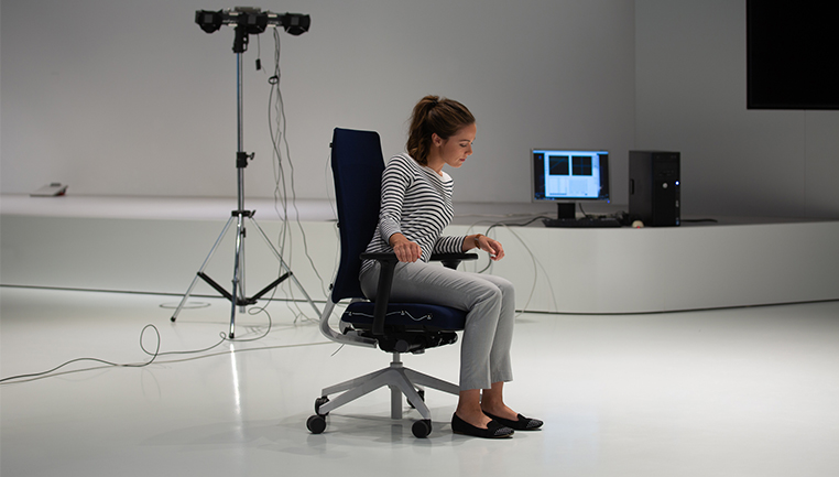 Una joven está sentada en la silla de escritorio Joyce en color azul oscuro con brazos en T en color negro y piezas de polipropileno en blanco. Gracias al sistema mecánico Flextech que favorece el movimiento en la silla giratoria de oficina Joyce, está sentada activamente.