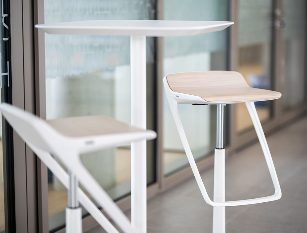 Close-up, two KINETIC bar stools with a white base frame and wooden seat surface in addition to a high white bistro table with a square table top.